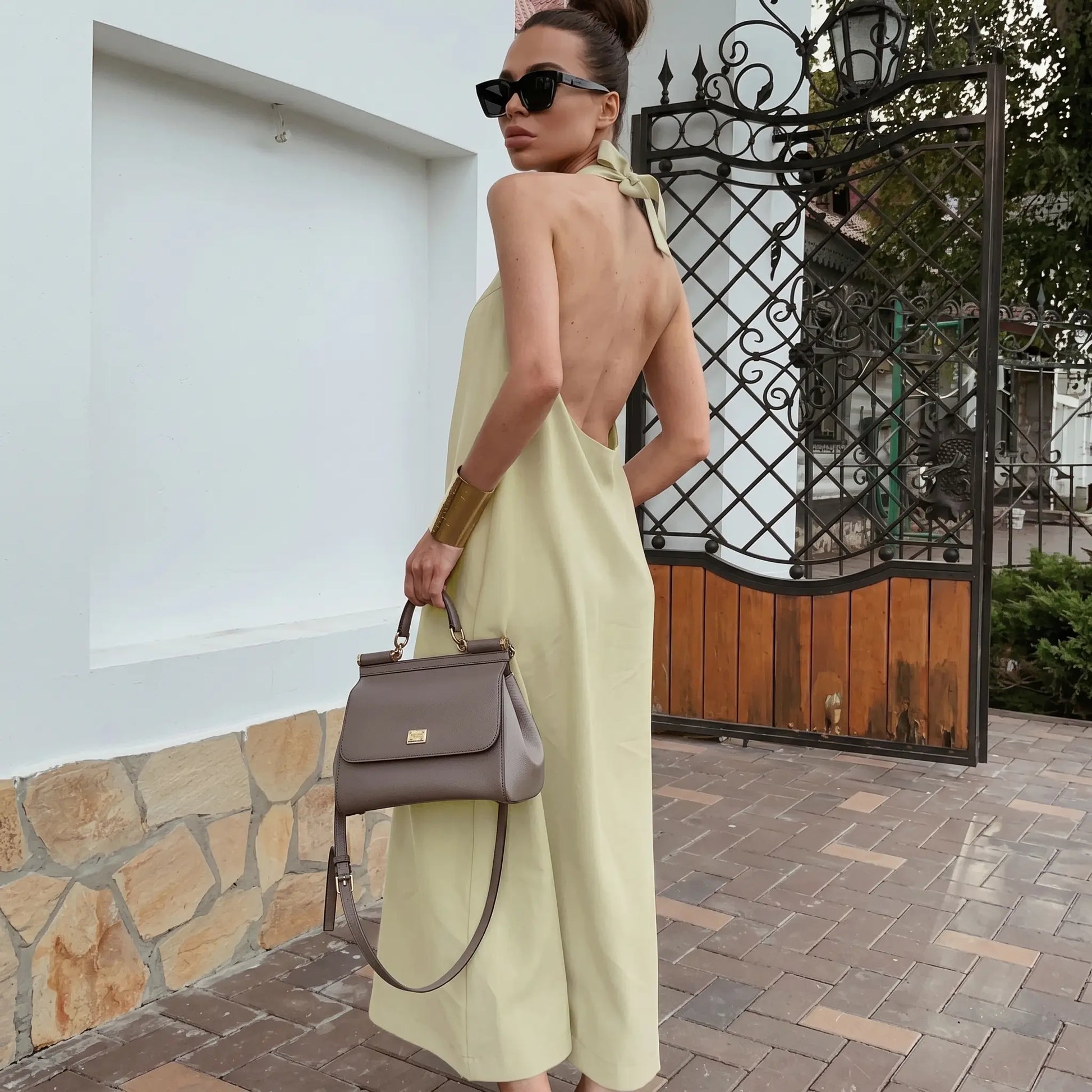 A woman models a flowing, light beige halter neck maxi dress made from a cotton linen blend, showcasing its open back and elegant drape in an outdoor setting.