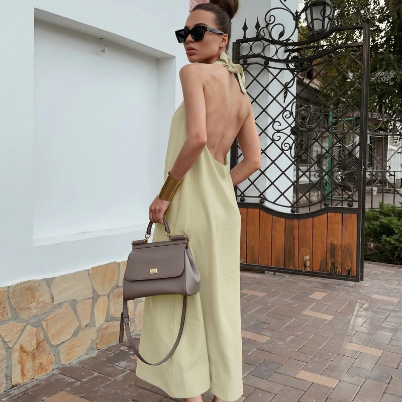 A woman models a flowing, light beige halter neck maxi dress made from a cotton linen blend, showcasing its open back and elegant drape in an outdoor setting.