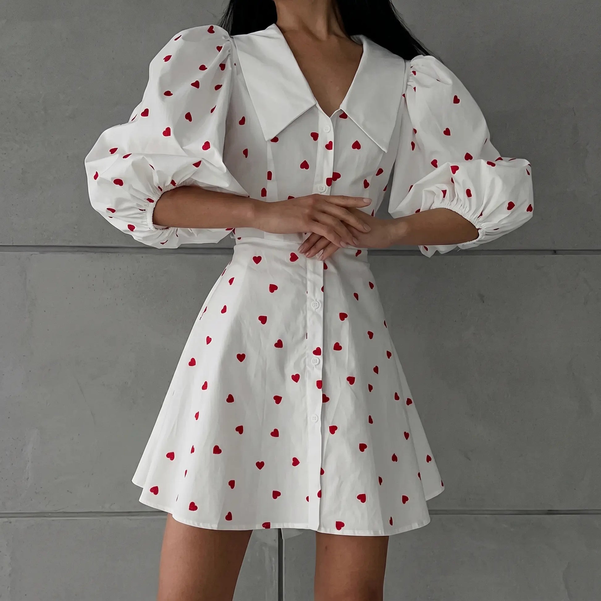 Woman wearing a white cotton linen blend A-line midi dress with a red heart print, featuring dramatic puff sleeves and a pointed collar, shown from the front.