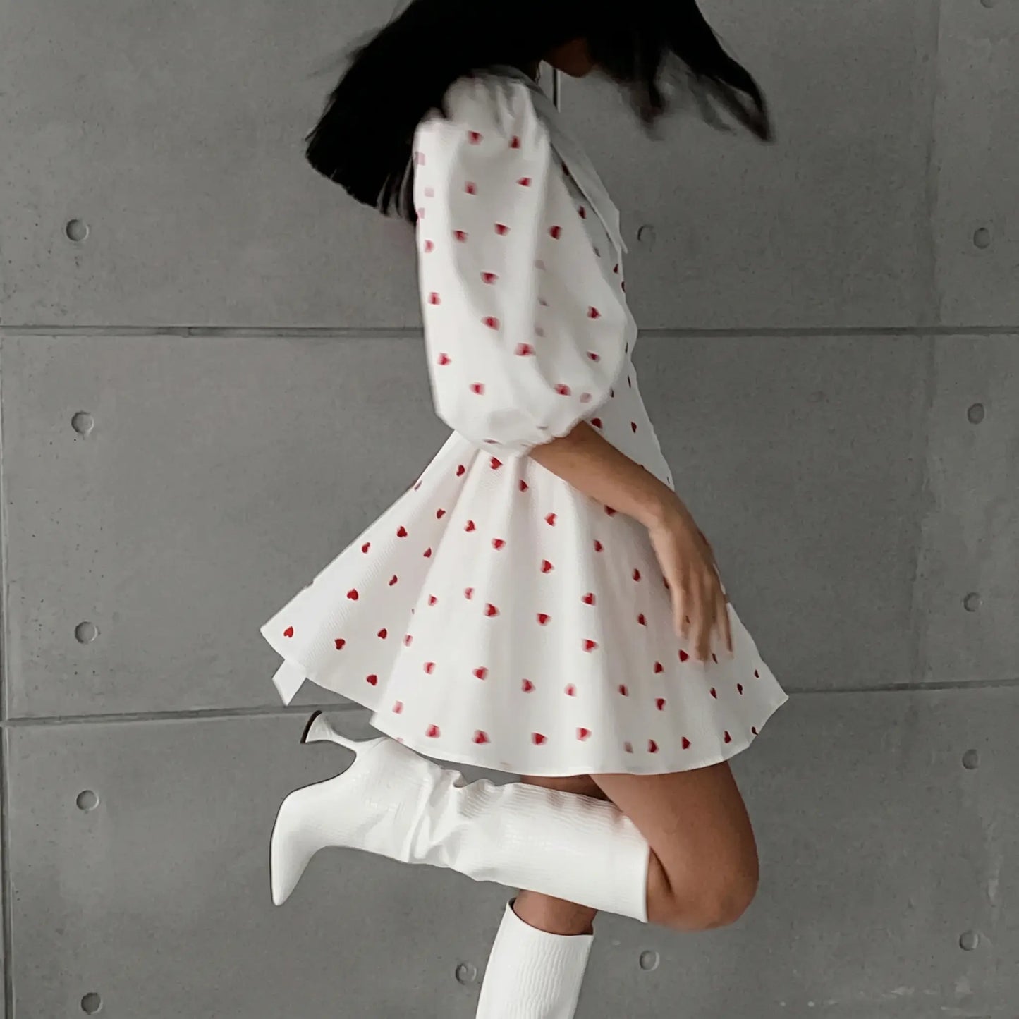 A woman in a white cotton-linen blend midi dress with a red heart print, featuring voluminous puff sleeves and a flared A-line skirt, styled with white boots.