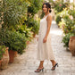Woman wearing a white midi dress and black patent stiletto heeled flip-flops, walking on a cobblestone path lined with lush plants in a Mediterranean courtyard.