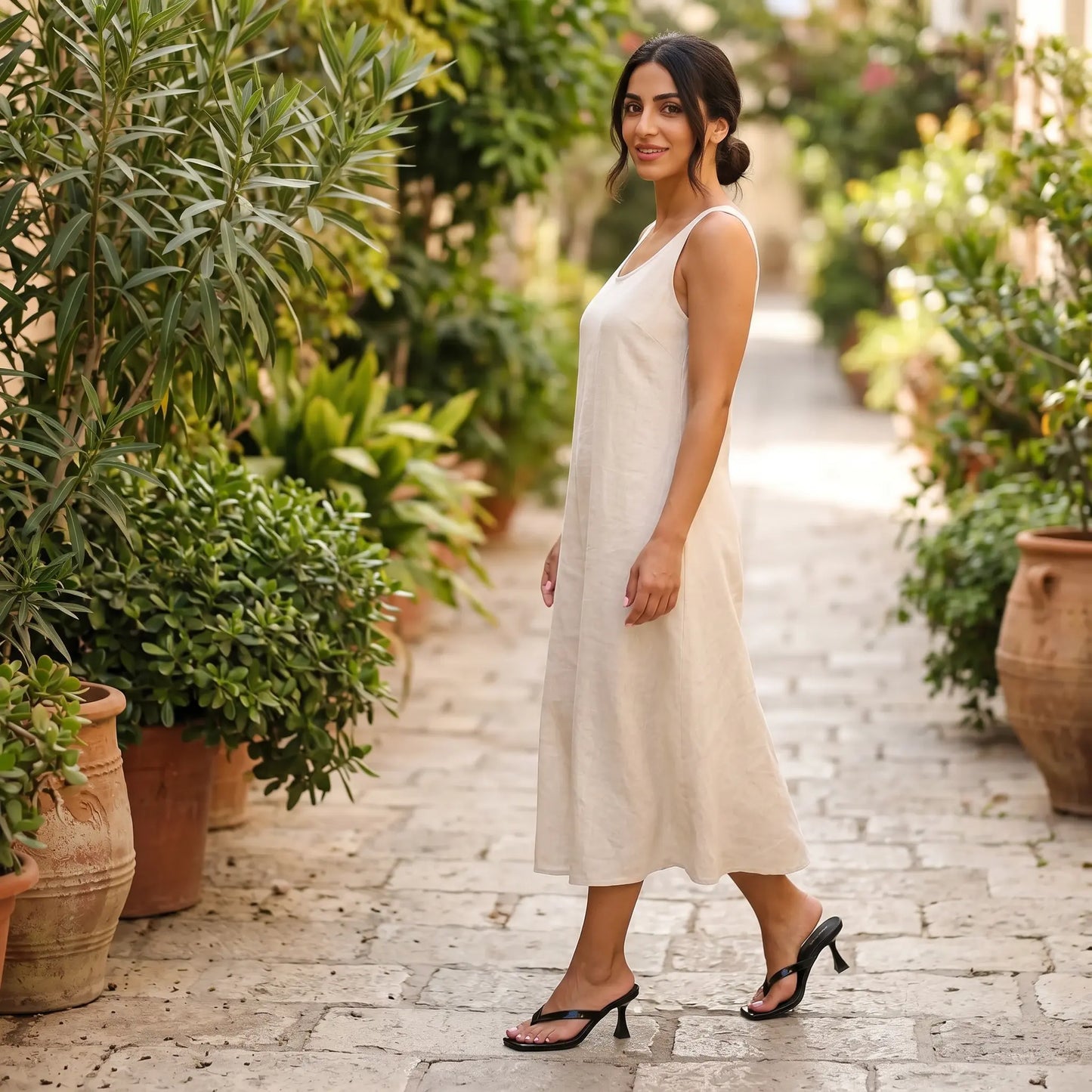 Woman wearing a white midi dress and black patent stiletto heeled flip-flops, walking on a cobblestone path lined with lush plants in a Mediterranean courtyard.