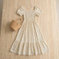 Ivory white polka dot maxi dress featuring a smocked square neck and short puff sleeves, styled flat-lay with a woven bag and pearl necklace on a textured beige surface.