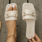 Jalani elegant ivory slide sandals shown on a foot with a gold anklet, featuring a square toe, embossed floral strap, and gold braided buckle, set against a weathered wooden background.