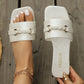Elegant white Jalani slide sandals with a square toe and embellished gold braided buckle, featuring an embossed floral pattern on the straps. Shown on a wooden surface with a model's foot adorned with a bracelet.