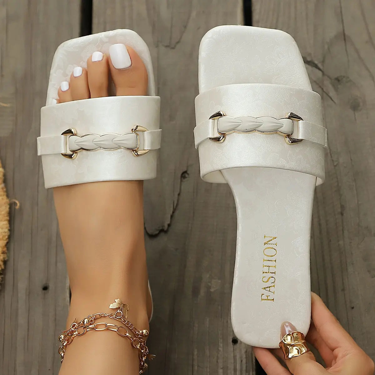 Elegant white Jalani slide sandals with a square toe and embellished gold braided buckle, featuring an embossed floral pattern on the straps. Shown on a wooden surface with a model's foot adorned with a bracelet.