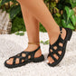 Elegant black matte synthetic leather strappy flat sandals with a gold-toned buckle, worn by a model on a decorative background of white pebbles and foliage. Features a rounded toe and secure ankle strap.