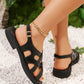 Black matte synthetic leather strappy flat sandals with a gold buckle, shown on a woman's feet in a lifestyle setting with white pebbles and greenery.