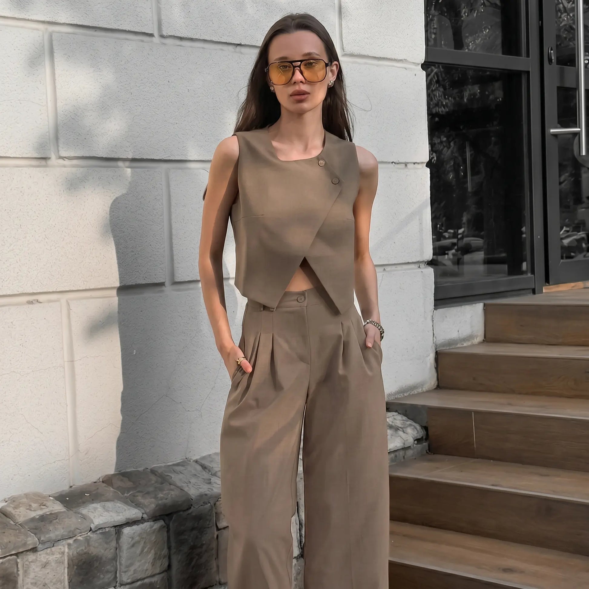 A woman models a chic beige asymmetrical crossover vest and high-waisted wide-leg trousers set in an outdoor lifestyle setting.