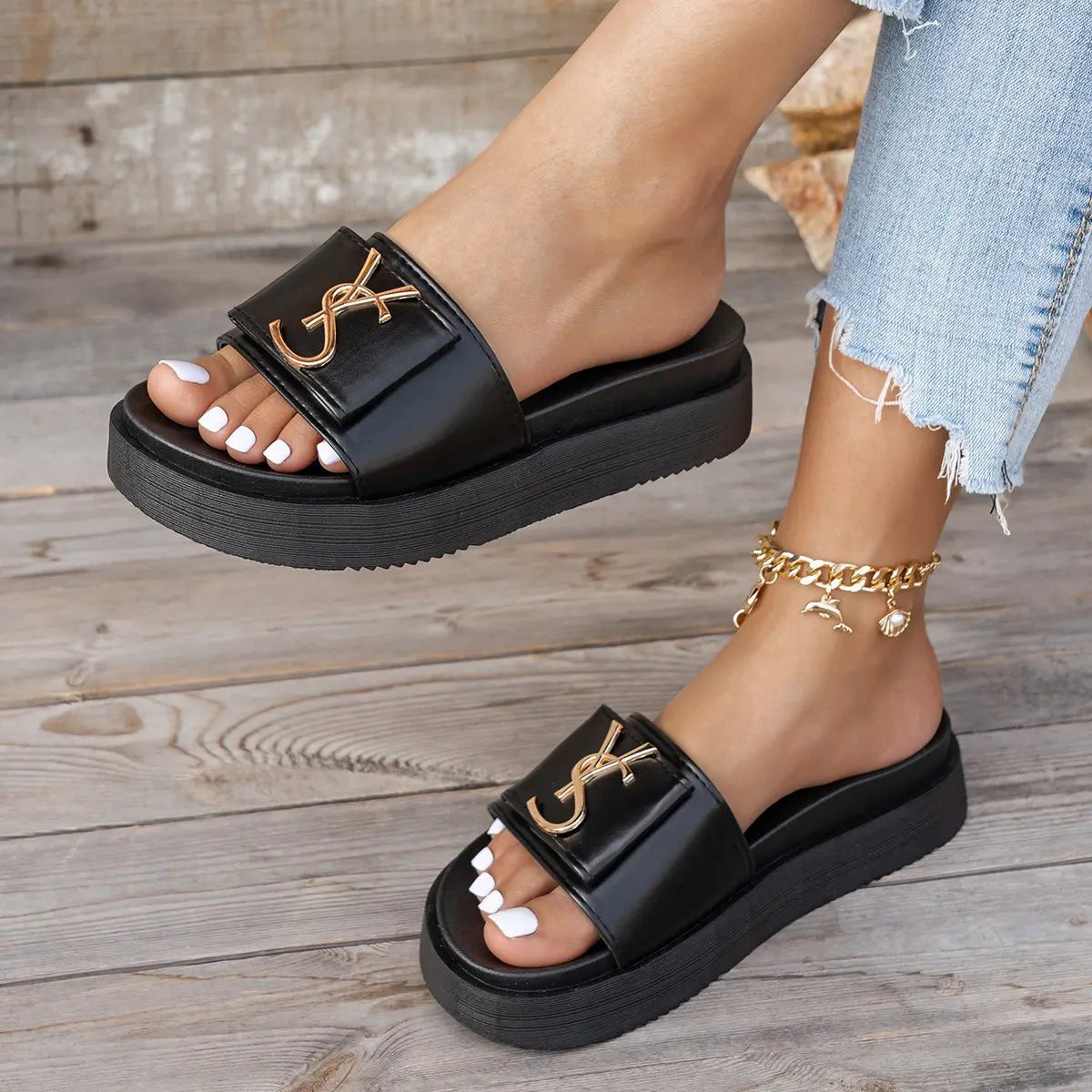 Black Kaelan platform slides with a wide strap and prominent polished gold metallic logo, worn casually with denim jeans and a gold anklet.