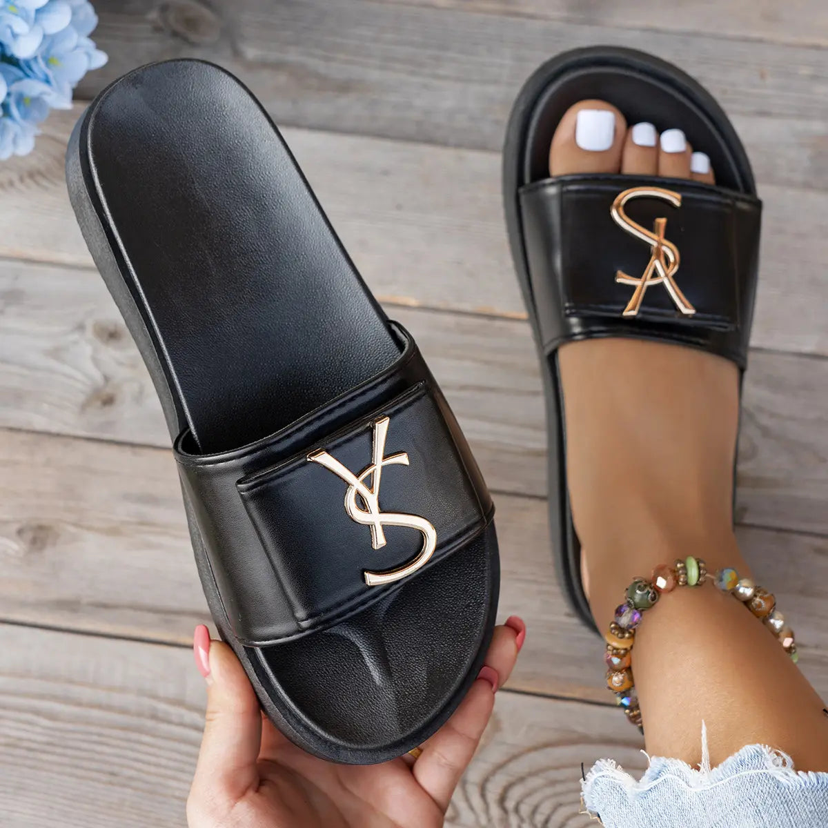 Black Kaelan platform slides featuring a wide strap with a polished gold metallic logo, worn on a foot with white nail polish and a beaded anklet, set against a rustic wooden surface.