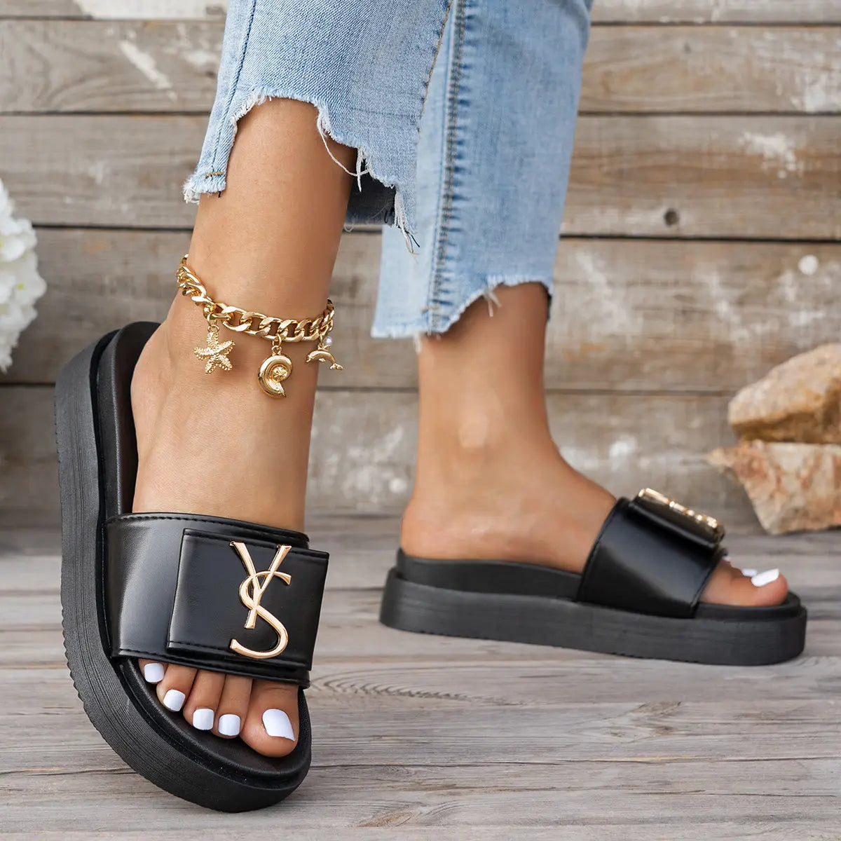 Close-up of black Kaelan platform slides with a wide vegan leather strap and polished gold-tone metallic logo, worn with distressed jeans and a charm anklet on a wooden deck.