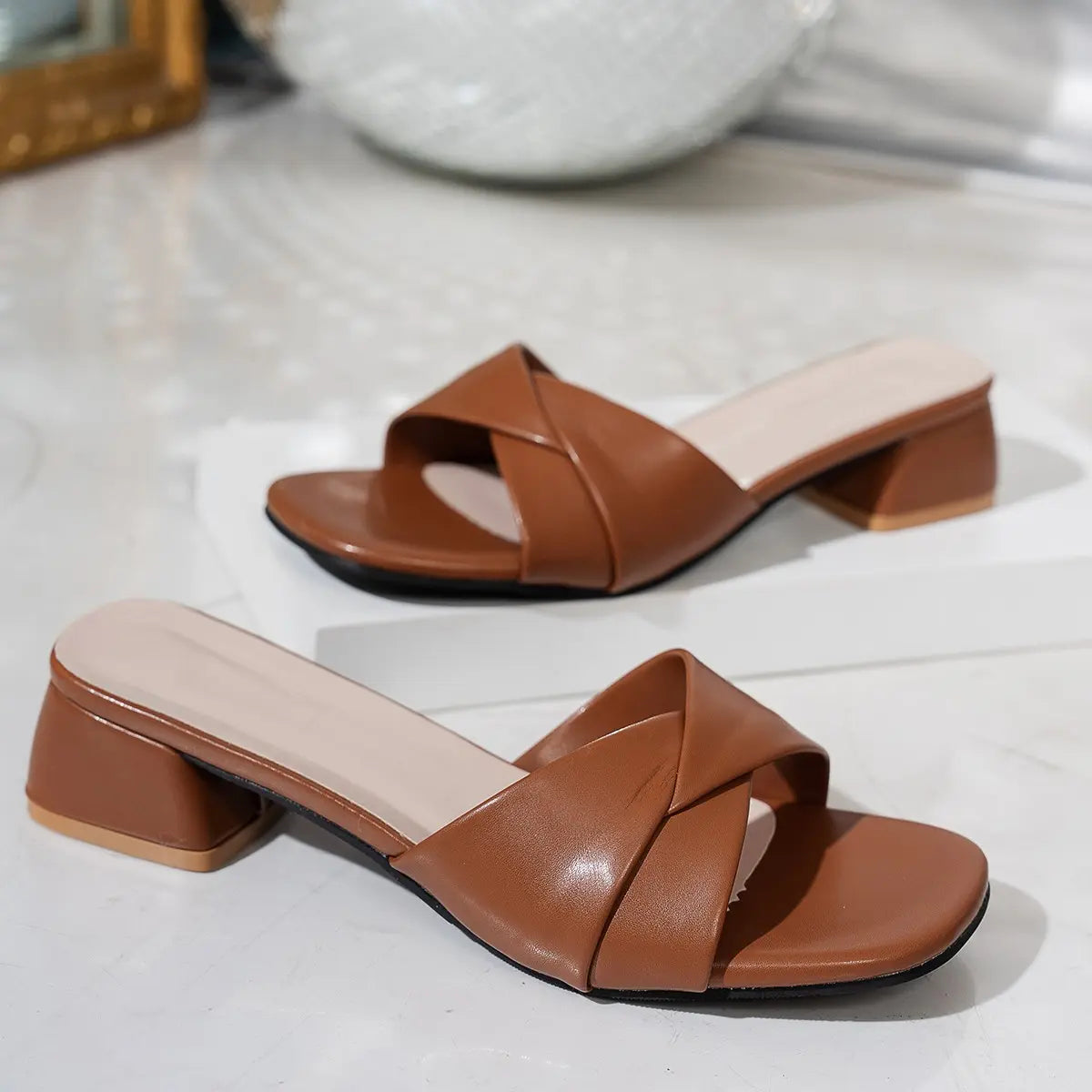 A pair of caramel brown Kaelan square-toe slide sandals with a chic twisted strap detail and a stable 4cm block heel, shown from a three-quarter front view on a studio background.