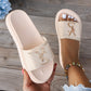 A pair of soft cream Kaelan platform slides worn on a foot, showcasing the wide strap with a polished gold-tone metallic logo accent and the thick, sculpted sole. Another slide is held in hand against a blurred wooden background.