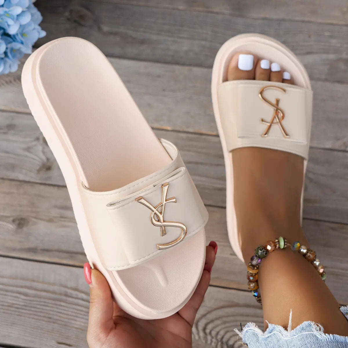 A pair of soft cream Kaelan platform slides worn on a foot, showcasing the wide strap with a polished gold-tone metallic logo accent and the thick, sculpted sole. Another slide is held in hand against a blurred wooden background.