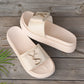 Cream Kaelan platform slide sandals with a wide PU leather strap featuring a polished gold-tone interlocking logo accent. Shown on a wooden surface with a palm leaf detail.