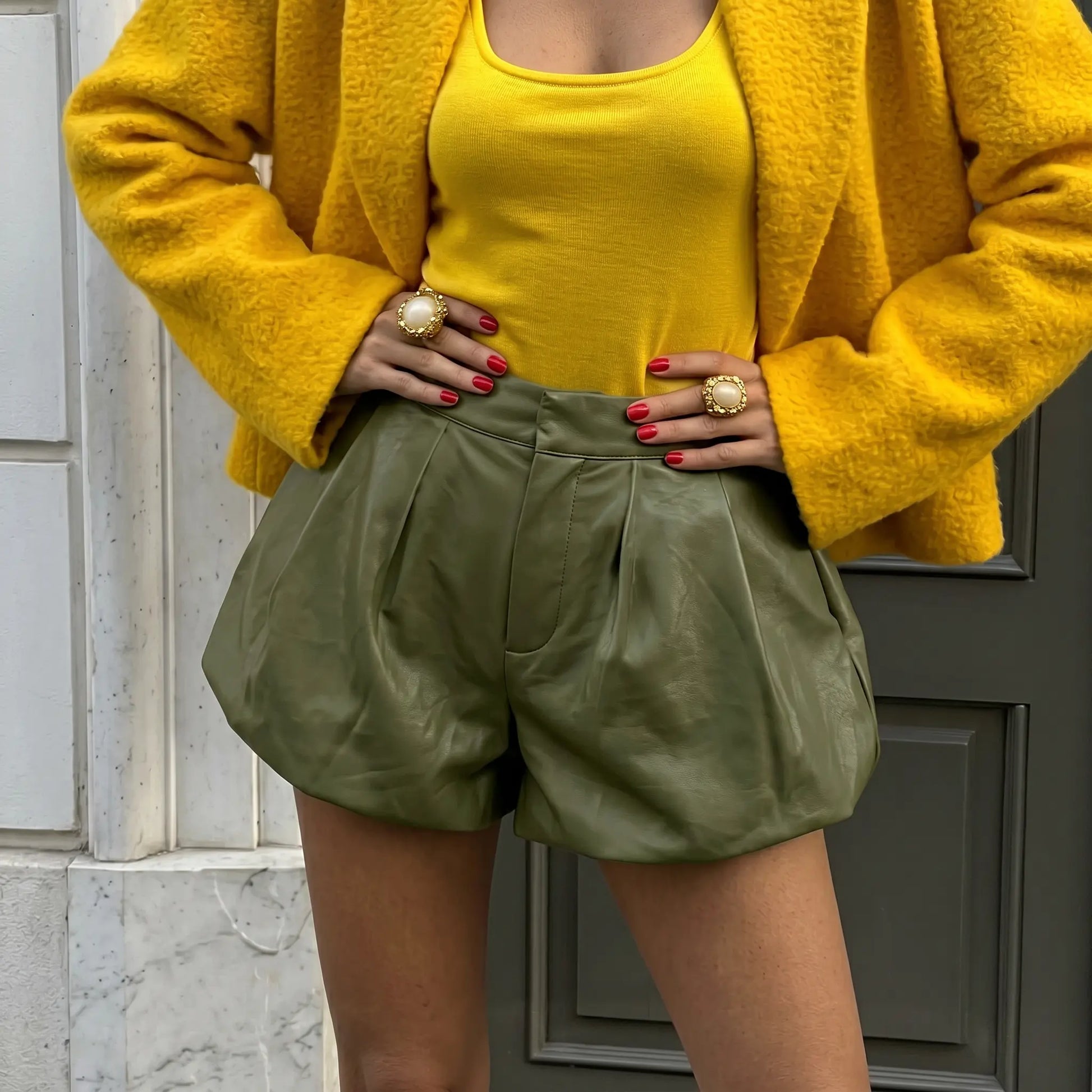 Olive green high-waist faux leather shorts with voluminous balloon silhouette and front pleats, styled with a yellow top and yellow blazer. These shorts showcase a chic, sophisticated bohemian look.