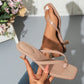 Elegant heeled thong sandals in apricot, worn on a foot with a squared toe and sculptural block heel, held by a hand against a white tiled background with flowers.