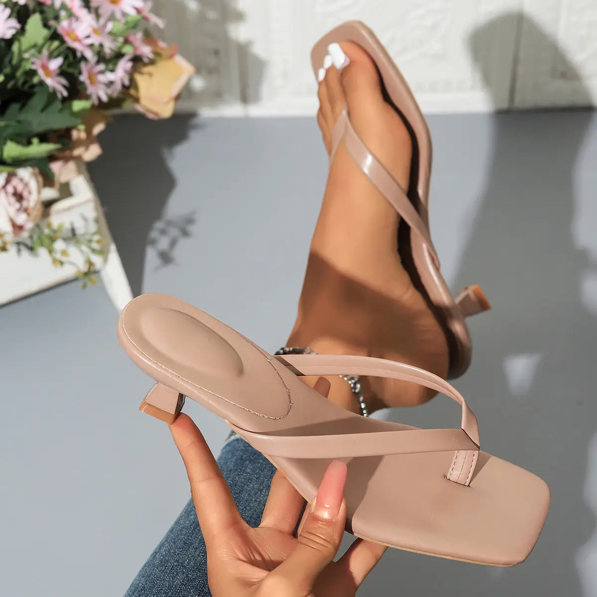 Elegant heeled thong sandals in apricot, worn on a foot with a squared toe and sculptural block heel, held by a hand against a white tiled background with flowers.