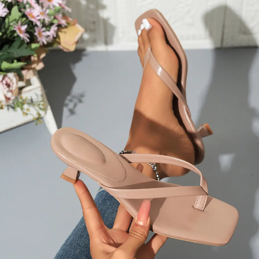 Elegant heeled thong sandals in apricot, worn on a foot with a squared toe and sculptural block heel, held by a hand against a white tiled background with flowers.
