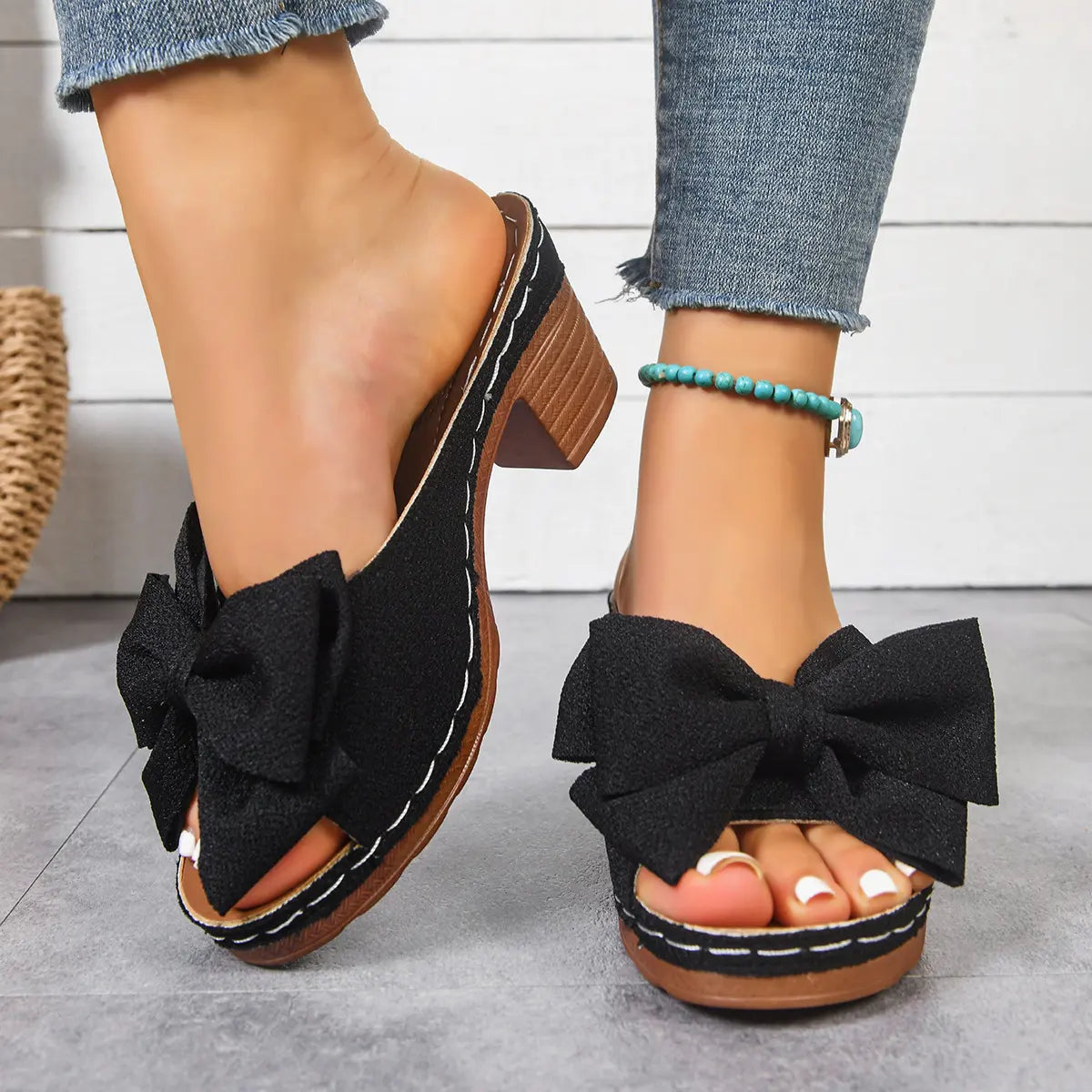 Black Kaelen women's mule sandals featuring a large textured fabric bow, fish-mouth peep-toe, and a 7cm wood-grain block heel with a 2cm platform, styled with blue jeans and a turquoise anklet.