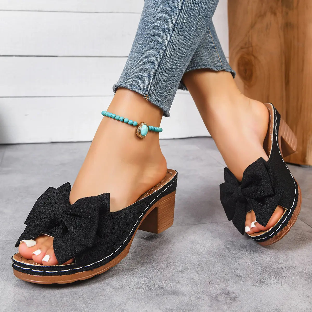 Women's black Kaelen fish-mouth mule sandals with a large textured bow detail, 7cm wood-grain block heel, and 2cm platform, worn with blue jeans and a turquoise anklet.