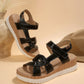 Kaelen women's black flat sandals featuring a multi-strap design with knot detail and white contrast stitching, resting on a wood-grain effect platform with adjustable hook-and-loop closures. A straw hat and decorative reeds complete the bohemian-inspired scene.