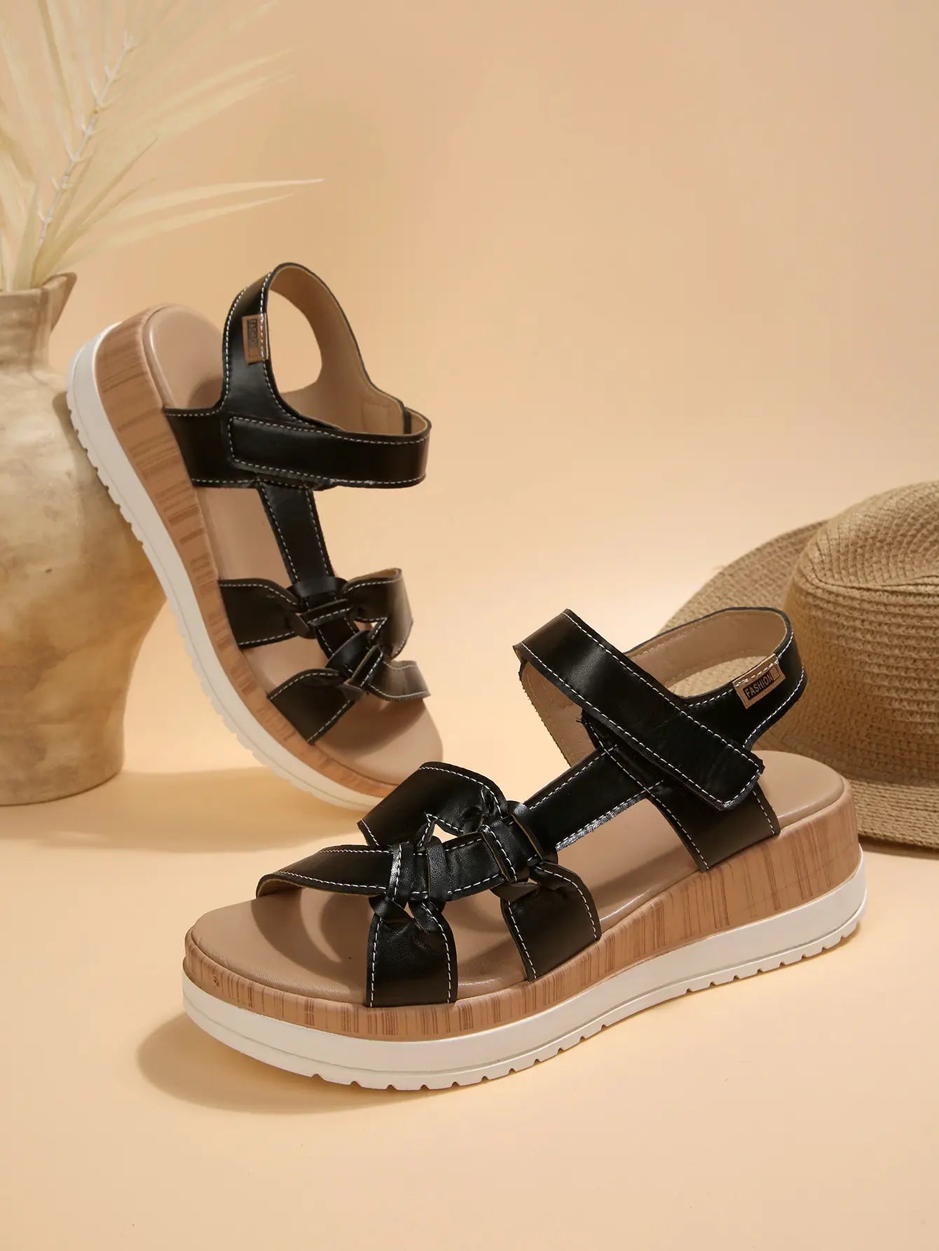 Kaelen women's black flat sandals featuring a multi-strap design with knot detail and white contrast stitching, resting on a wood-grain effect platform with adjustable hook-and-loop closures. A straw hat and decorative reeds complete the bohemian-inspired scene.