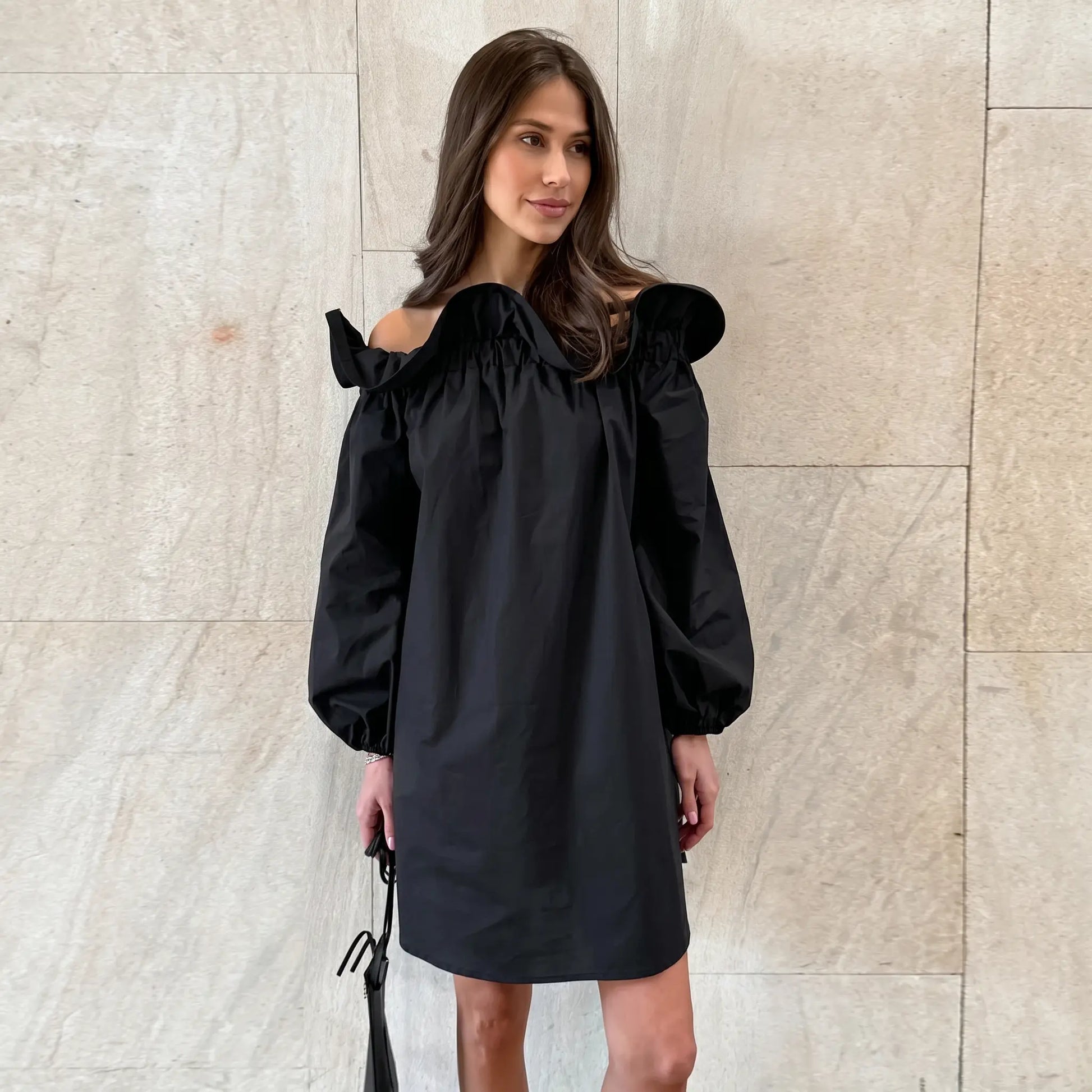 A Kaelen black off-shoulder cotton mini dress with romantic ruffled neckline and voluminous balloon sleeves, worn by a model against a textured stone wall.