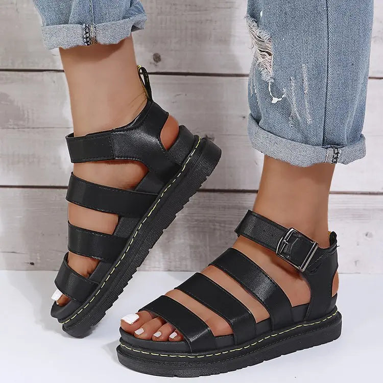 Black Kaelen platform gladiator sandals with chunky sole and black buckle strap, shown on a woman's feet with rolled jeans.