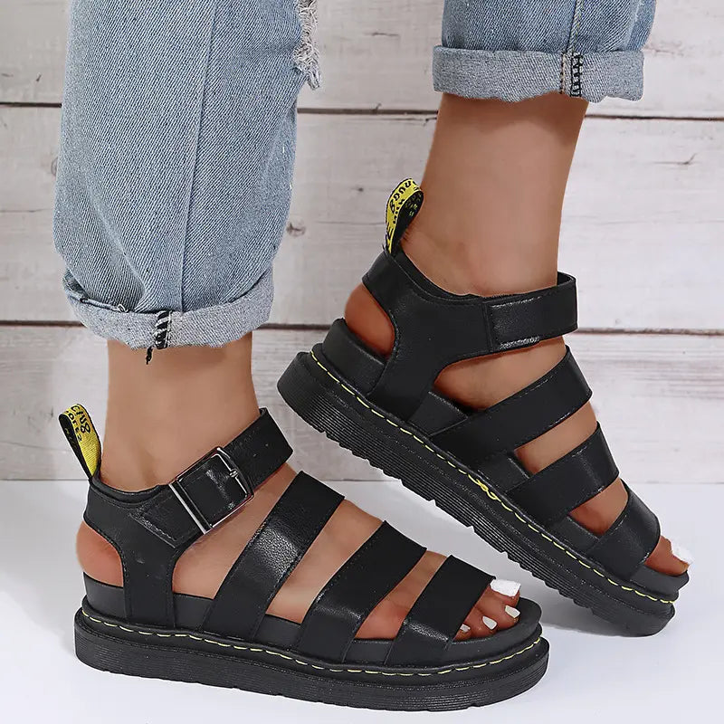Black Kaelen platform gladiator sandals shown on a person's feet, featuring a chunky 3cm sole with yellow stitching detail and a black buckle ankle strap.