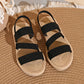 Black woven strap flat sandals with a braided sole trim, shown from a top-down view on a woven mat background.