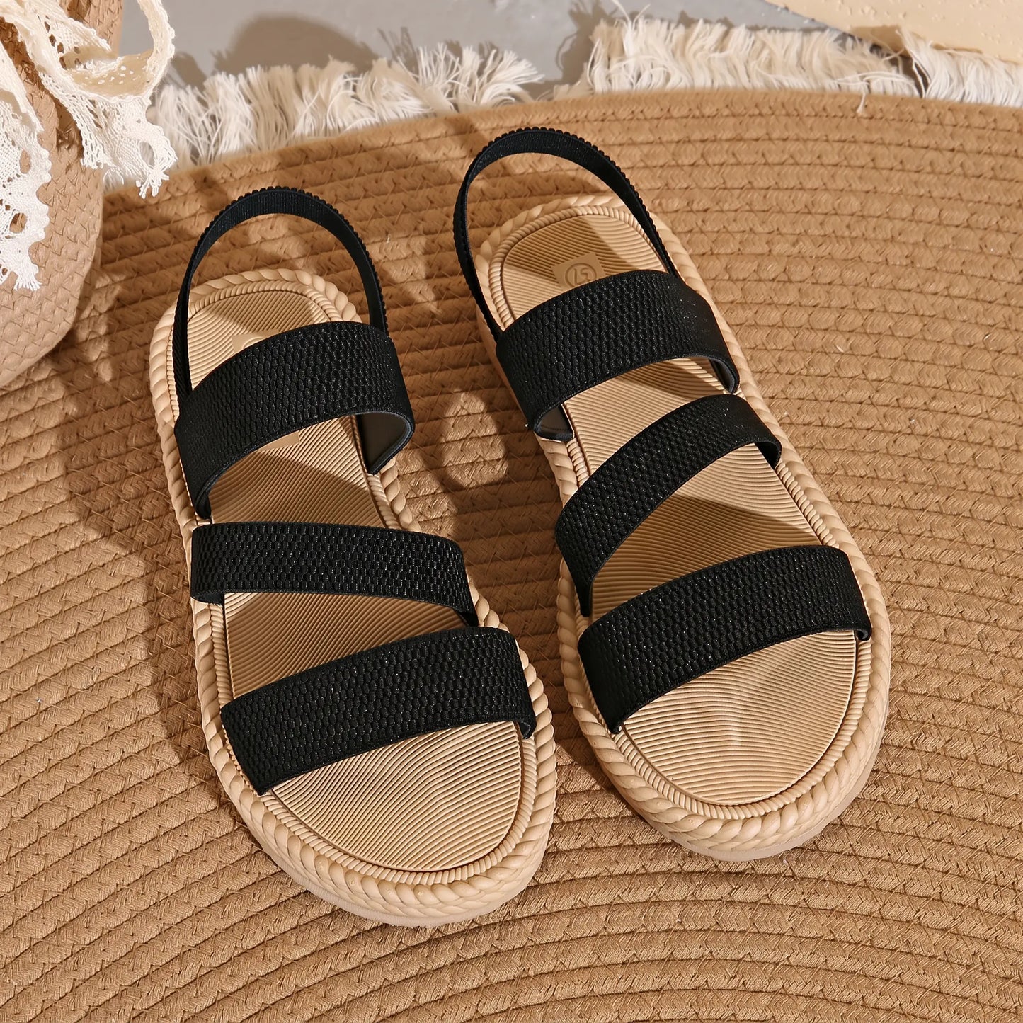 Black woven strap flat sandals with a braided sole trim, shown from a top-down view on a woven mat background.