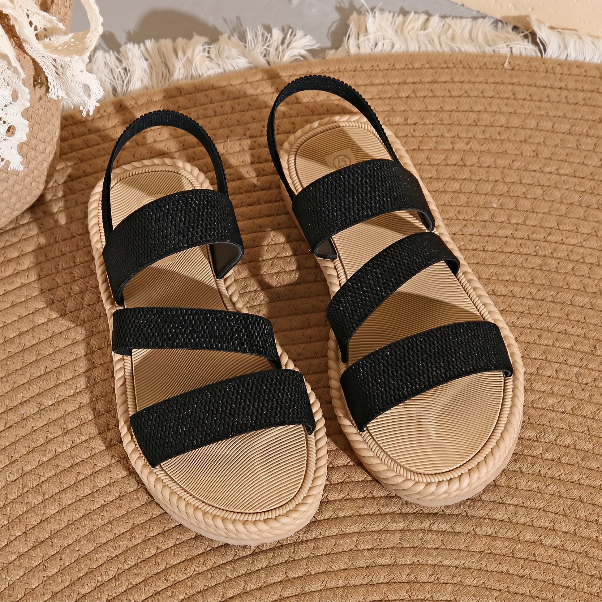Black woven strap flat sandals with a braided sole trim, shown from a top-down view on a woven mat background.