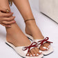 Elegant off-white patent Kaelen flat sandals featuring a prominent dark red bow embellishment, shown on a model's feet with a clean studio background.