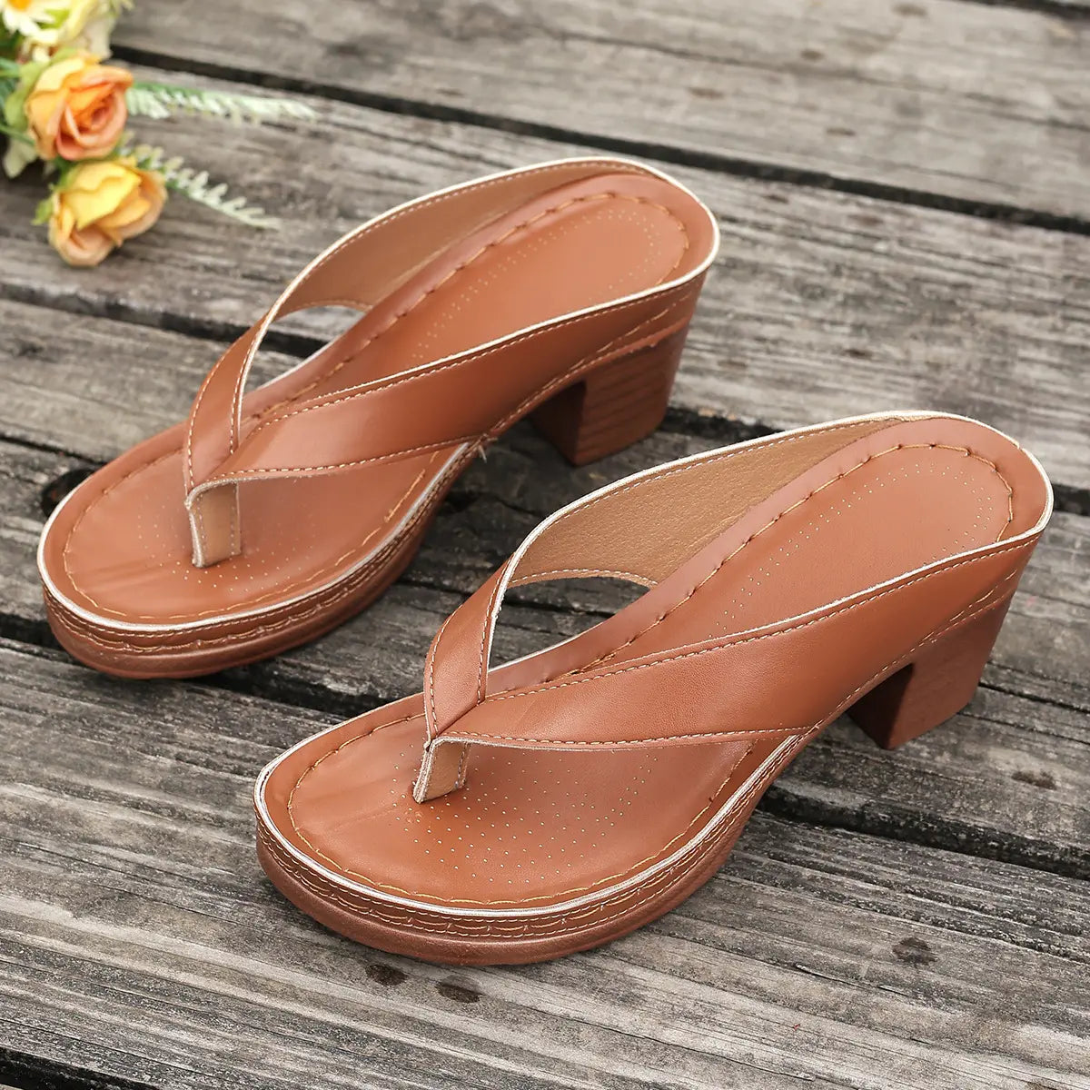 Brown PU leather heeled slides with thong strap and platform sole, shown at a three-quarter angle on a rustic wooden surface with yellow roses.