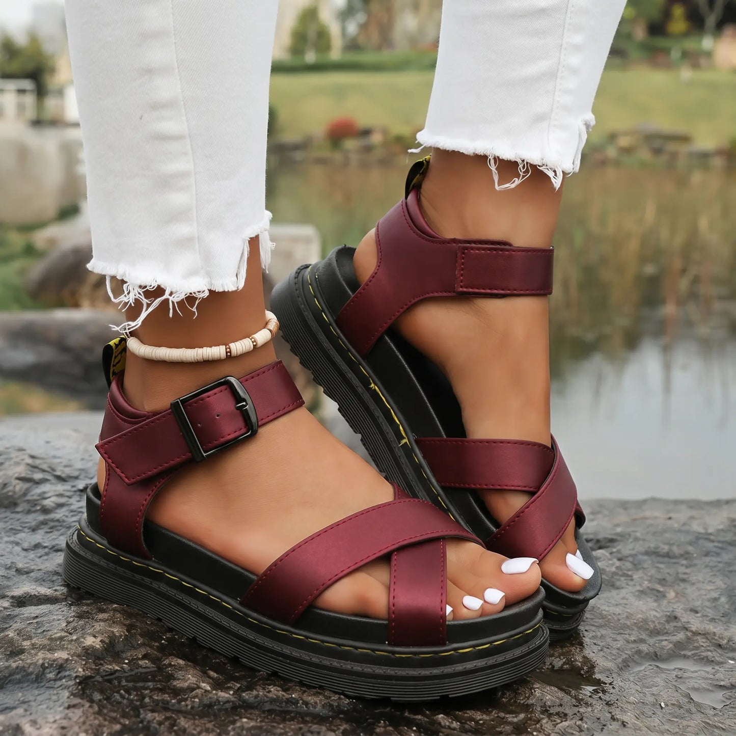 Deep wine red Kaelen platform sandals featuring criss-cross straps and a chunky lugged sole, worn by a model with ripped white jeans on natural rock formations.