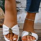 A woman's feet wearing Kaelen woven flat slide sandals in ivory weave, featuring a textured strap and a gold oval buckle, paired with distressed denim jeans. The sandals rest on a wooden surface, evoking a bohemian summer style.