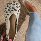 A pair of women's Kaelen knit ballet flats in leopard print, shown being worn on a foot and held by a hand, styled with distressed denim jeans on a woven mat.