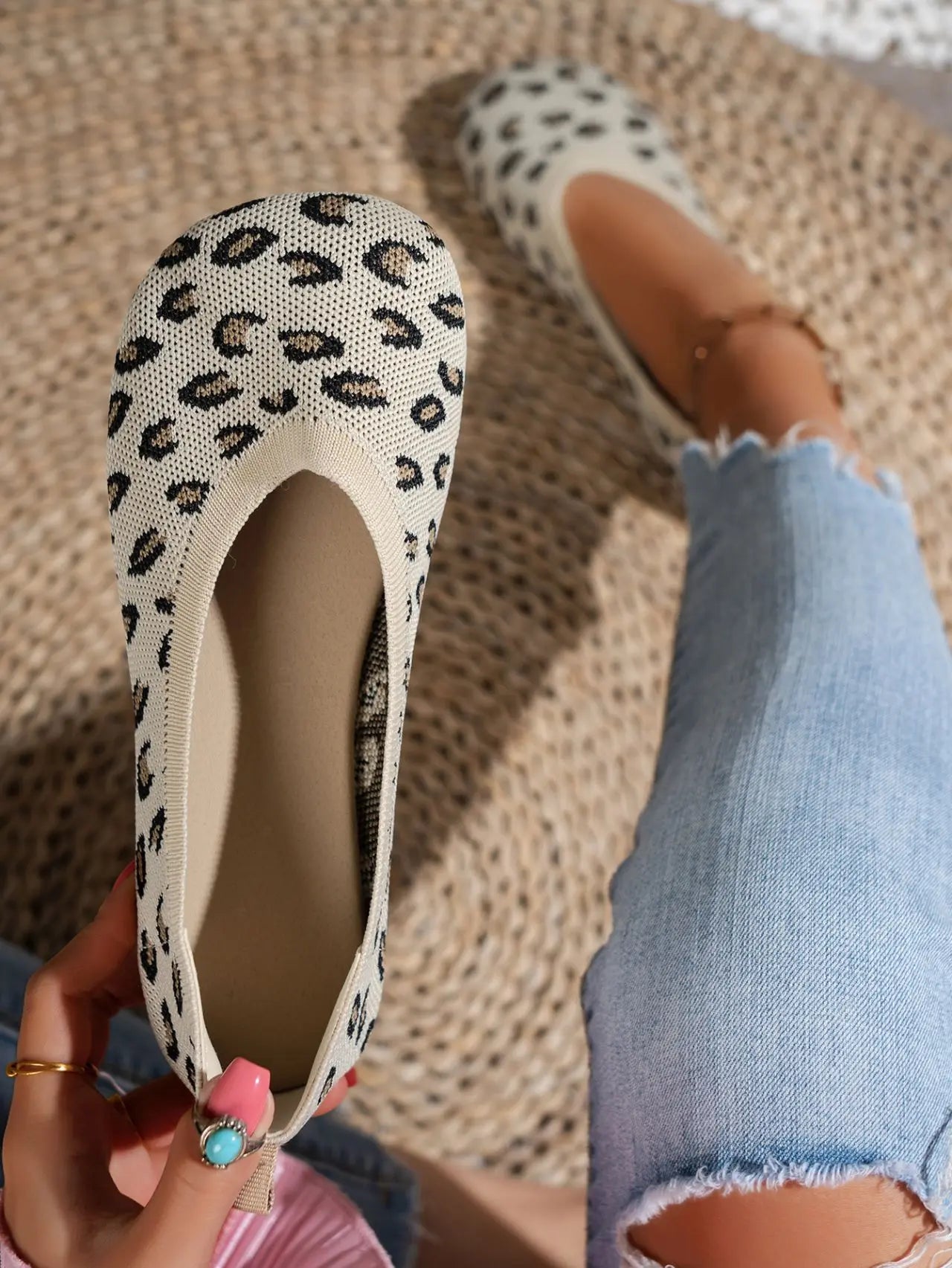 A pair of women's Kaelen knit ballet flats in leopard print, shown being worn on a foot and held by a hand, styled with distressed denim jeans on a woven mat.