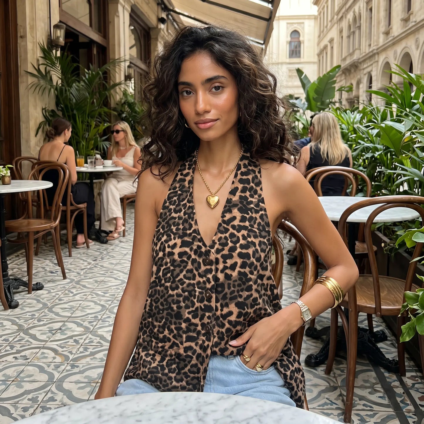 Woman wearing a Kaelen leopard print halter neck top and light blue jeans, sitting at an outdoor cafe table with a gold heart pendant necklace.