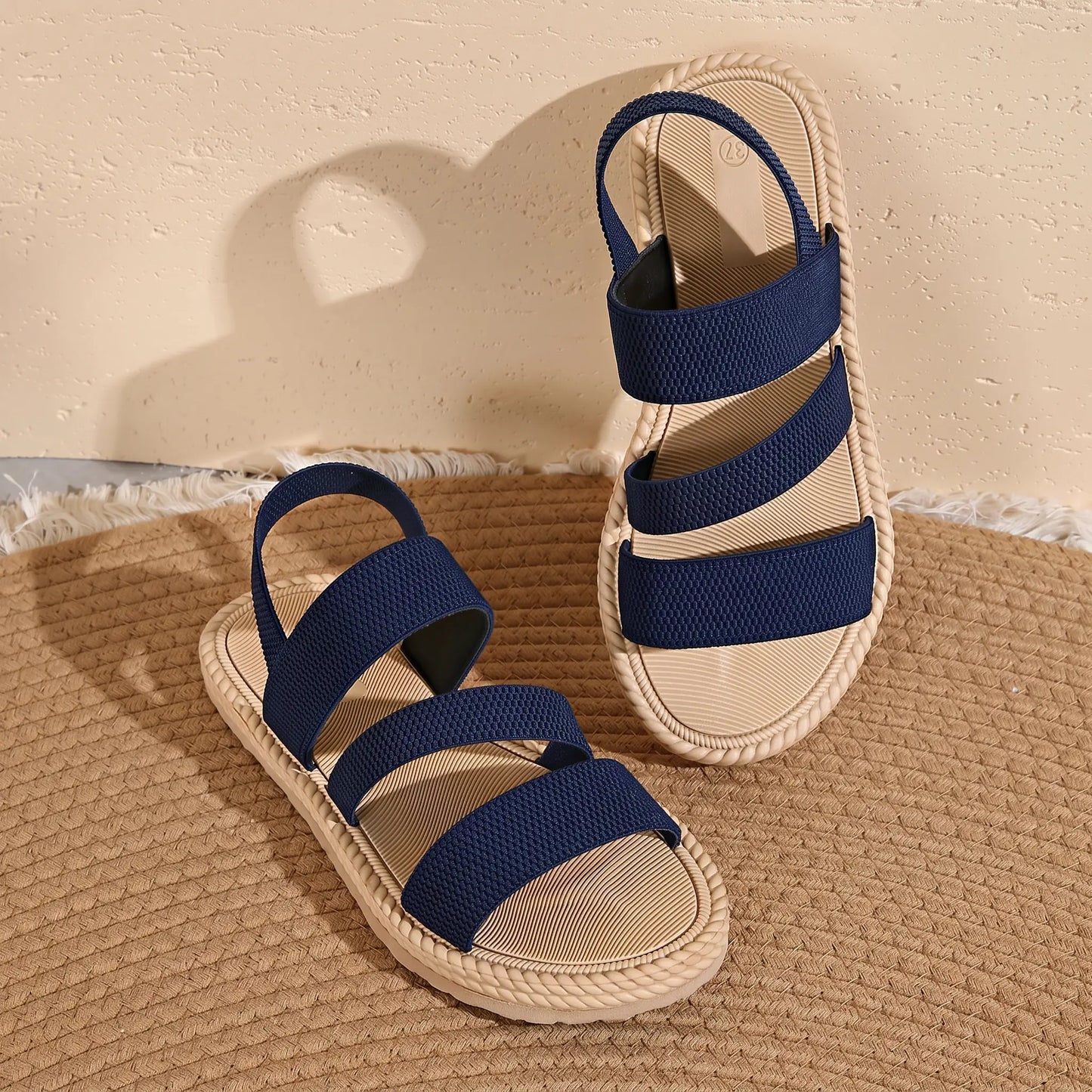 Navy blue flat Kaelen sandals featuring three parallel woven elastic straps and a rear heel strap, presented on a woven mat with a textured background. Side view showcases the comfortable, bohemian-chic design.
