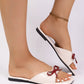 Kaelen women's off-white patent flat sandals featuring a prominent dark red bow embellishment and an open square toe, worn on a model's feet with a gold anklet. These chic slip-on summer sandals offer comfortable, elegant style.