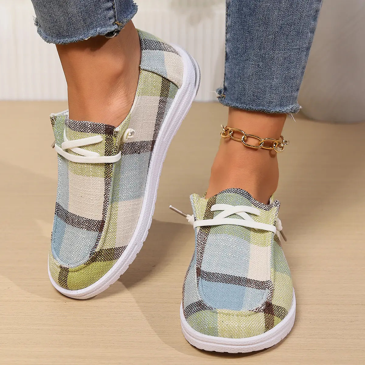 Women's Kaelen plaid canvas slip-on loafers in a green, blue, and beige pattern, styled with denim jeans and a gold anklet. Round toe casual flats with white laces and flat white rubber soles.
