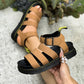 Close-up view of tan faux leather platform gladiator sandals on a foot, showcasing the chunky black sole with yellow stitching and an adjustable ankle strap. A second sandal is held by a hand with a pearl ring, against a blurred background of concrete and greenery.