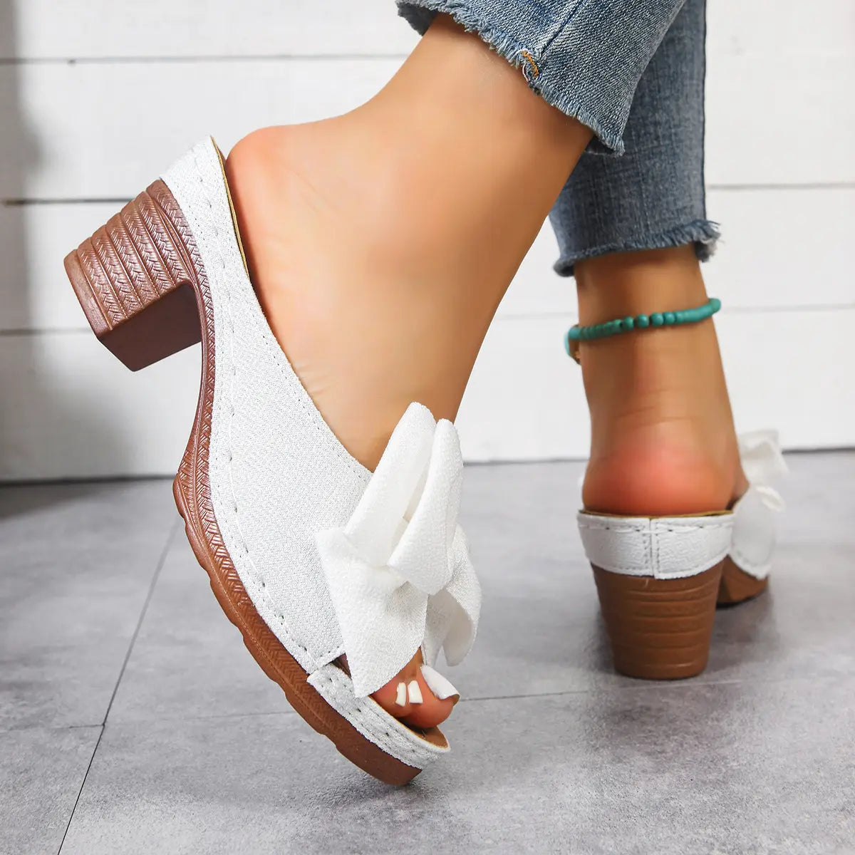 Women's Kaelen white slip-on mule sandals with textured fabric bow detail, fish-mouth peep-toe, and a 7cm wood-grain block heel worn with blue denim jeans and a turquoise anklet.