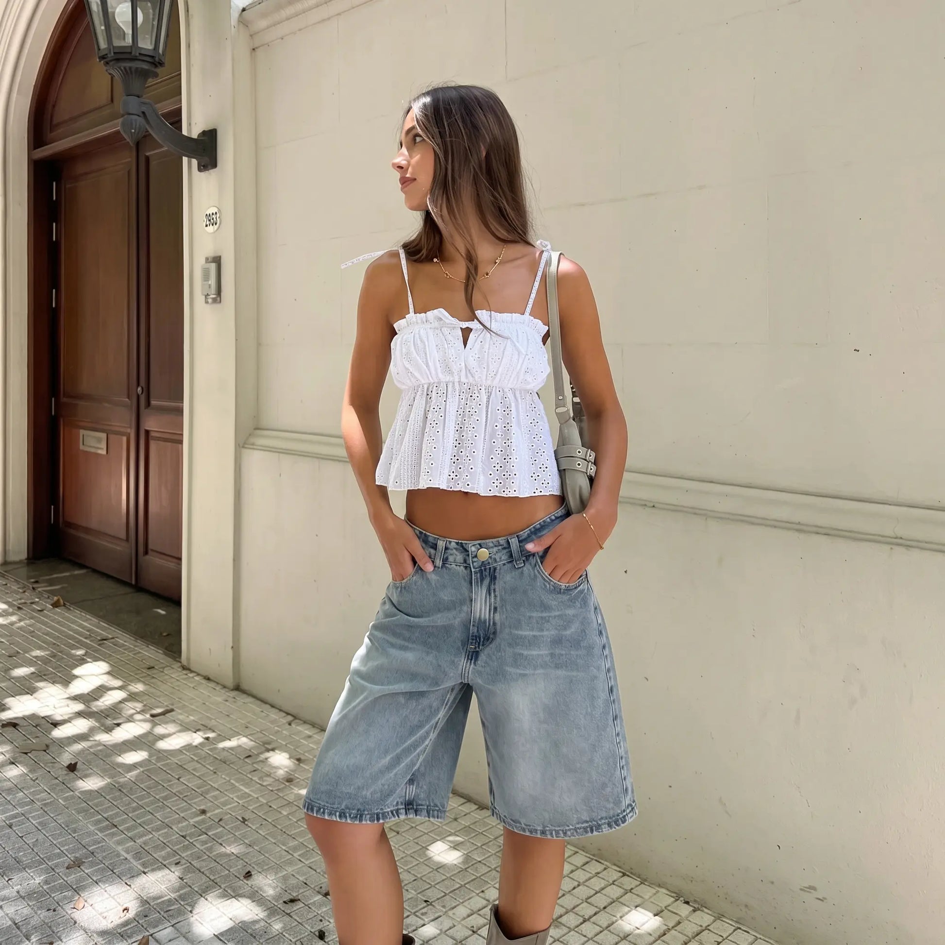 Model wears a white broderie anglaise camisole top with a ruffled, tie-detail neckline and adjustable shoulder straps, styled with denim shorts.