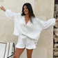 Woman wearing a white linen-blend set featuring a loose V-neck blouse with voluminous lantern sleeves and high-waisted shorts with a delicate ruffled hem, styled in a bohemian summer setting.