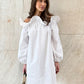 Kaelen white off-shoulder cotton mini dress with voluminous balloon sleeves, worn by a model holding a black handbag against a textured wall.