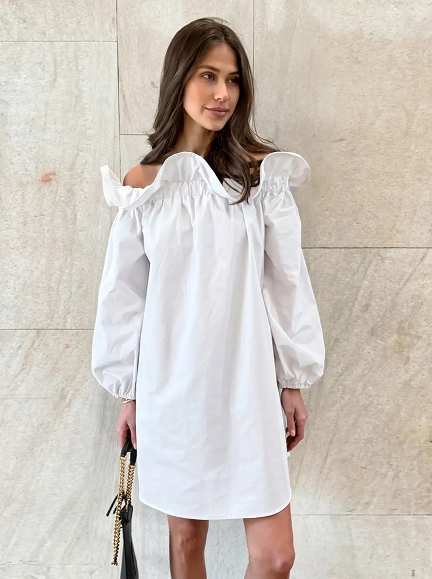 Kaelen white off-shoulder cotton mini dress with voluminous balloon sleeves, worn by a model holding a black handbag against a textured wall.